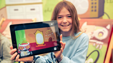 Mädchen hält ein Tablet mit laufendem KatzeQ-Spiel und lächelt in die Kamera, Release-Pressefoto.